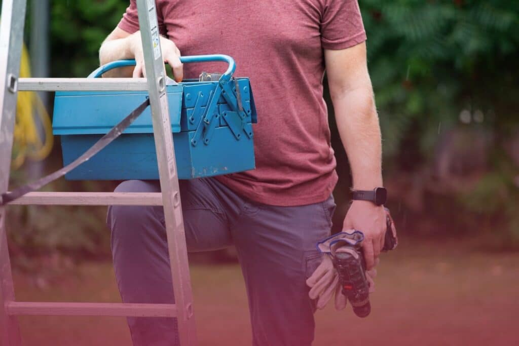 male homeowner holds tools and is resting his leg on a ladder while doing home maintenance