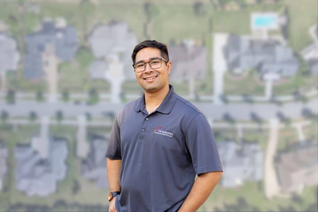 A smiling man in a branded grey home inspector polo shirt standing against a blurred aerial backdrop of a residential neighborhood.