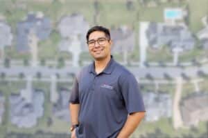 A smiling man in a branded grey home inspector polo shirt standing against a blurred aerial backdrop of a residential neighborhood.