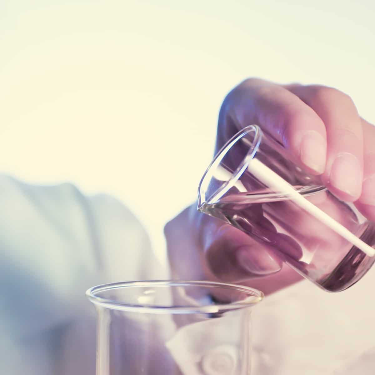 A scientist's hand in a lab coat tilts a small beaker to pour a clear liquid into a larger beaker.