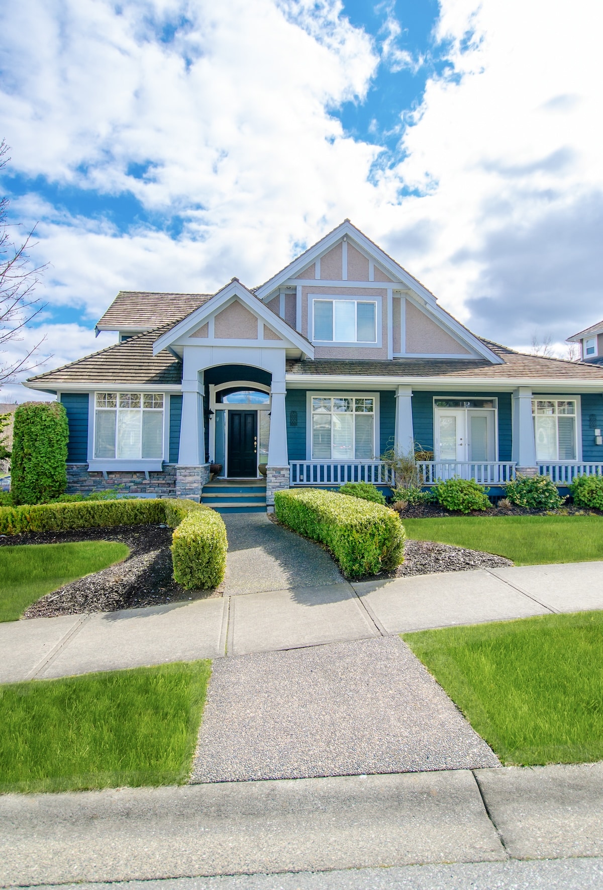 The front exterior of a two-story suburban house featuring dark blue siding, light gray gables, stone-accented pillars, and a covered porch. A paved walkway leads to the black front door, flanked by a neatly manicured green lawn and trimmed hedges under a bright blue sky with white clouds.