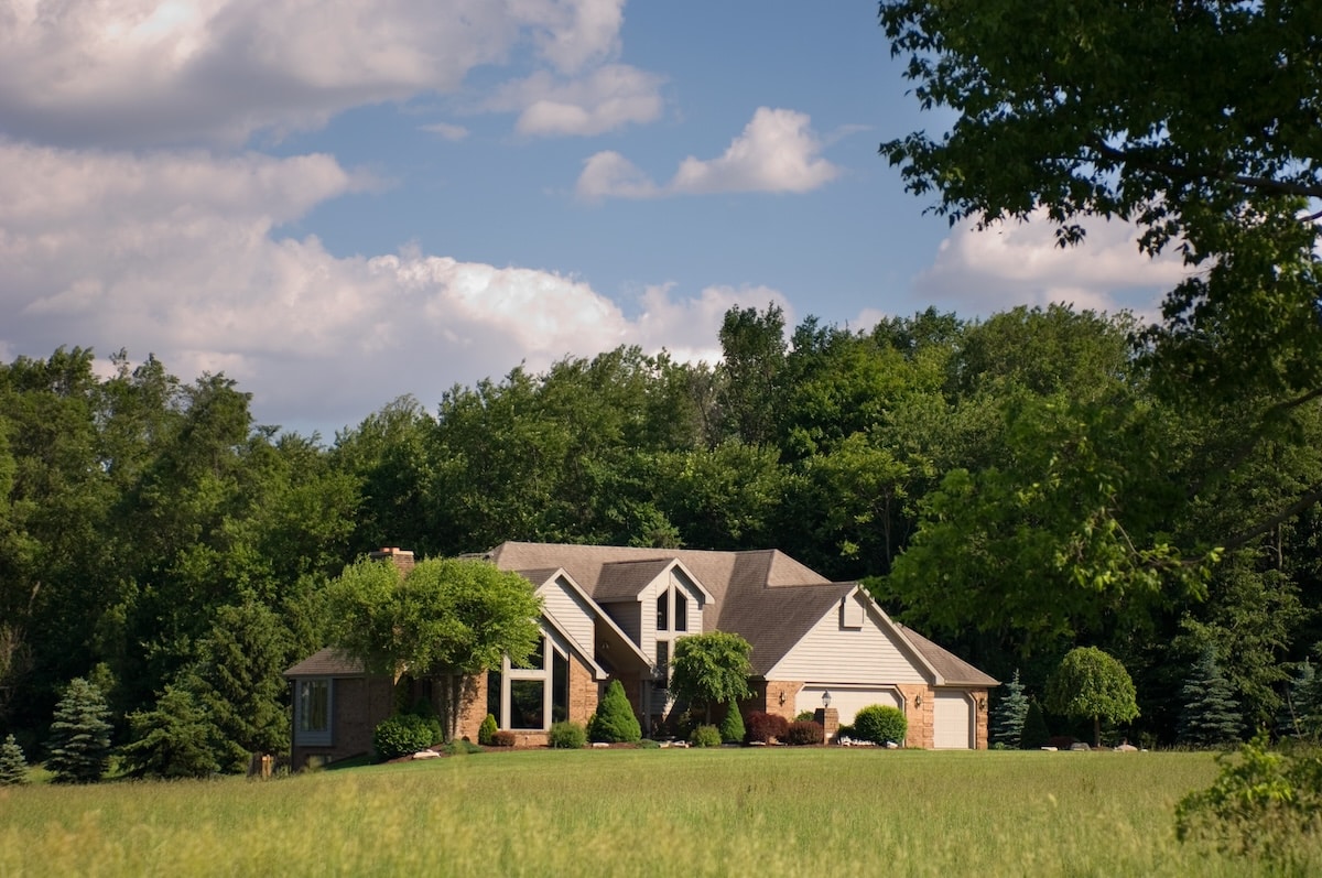 A large, multi-gabled suburban home with light beige siding, brick accents, and a two-car garage, situated in front of a dense green forest and a wide grassy field.