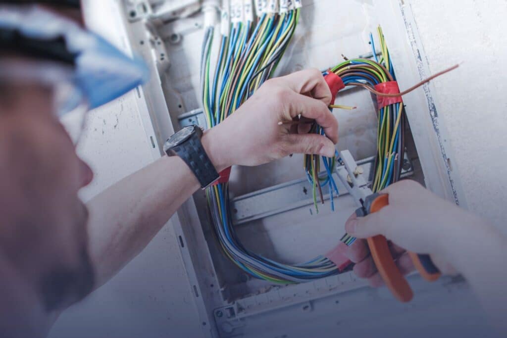Close-up of an electrician using pliers to strip and organize colorful electrical wires inside an open circuit breaker panel.
