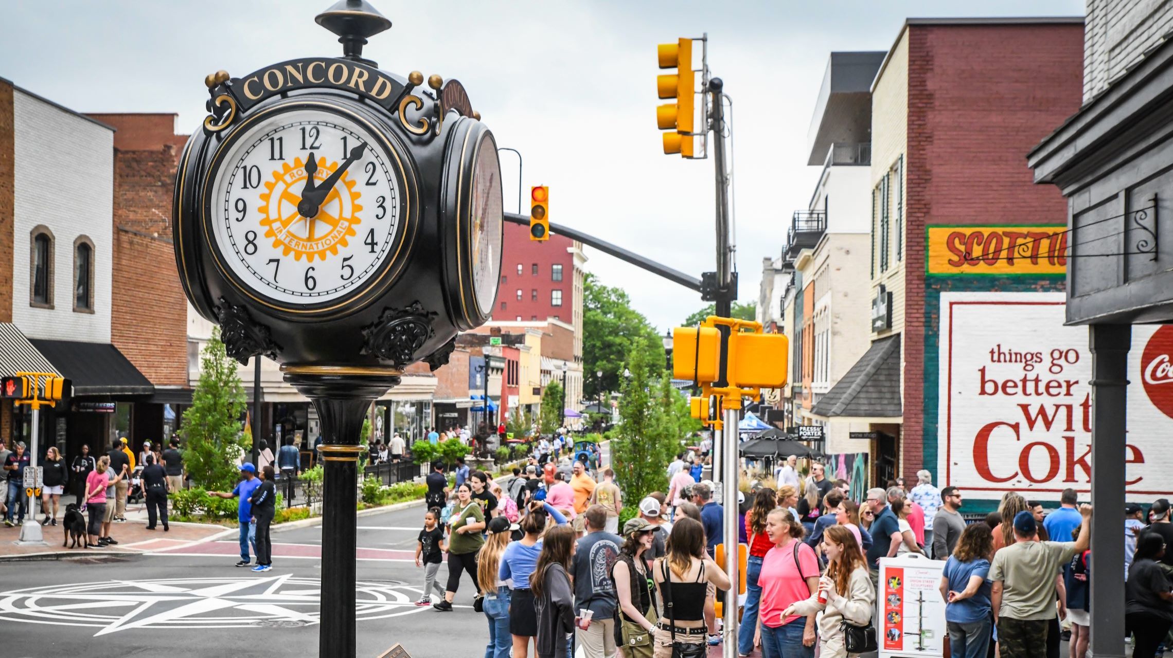A large black vintage street clock labeled "Concord" standing in the foreground on a bustling downtown street filled with pedestrians, storefronts, and a vintage Coca-Cola wall sign.
