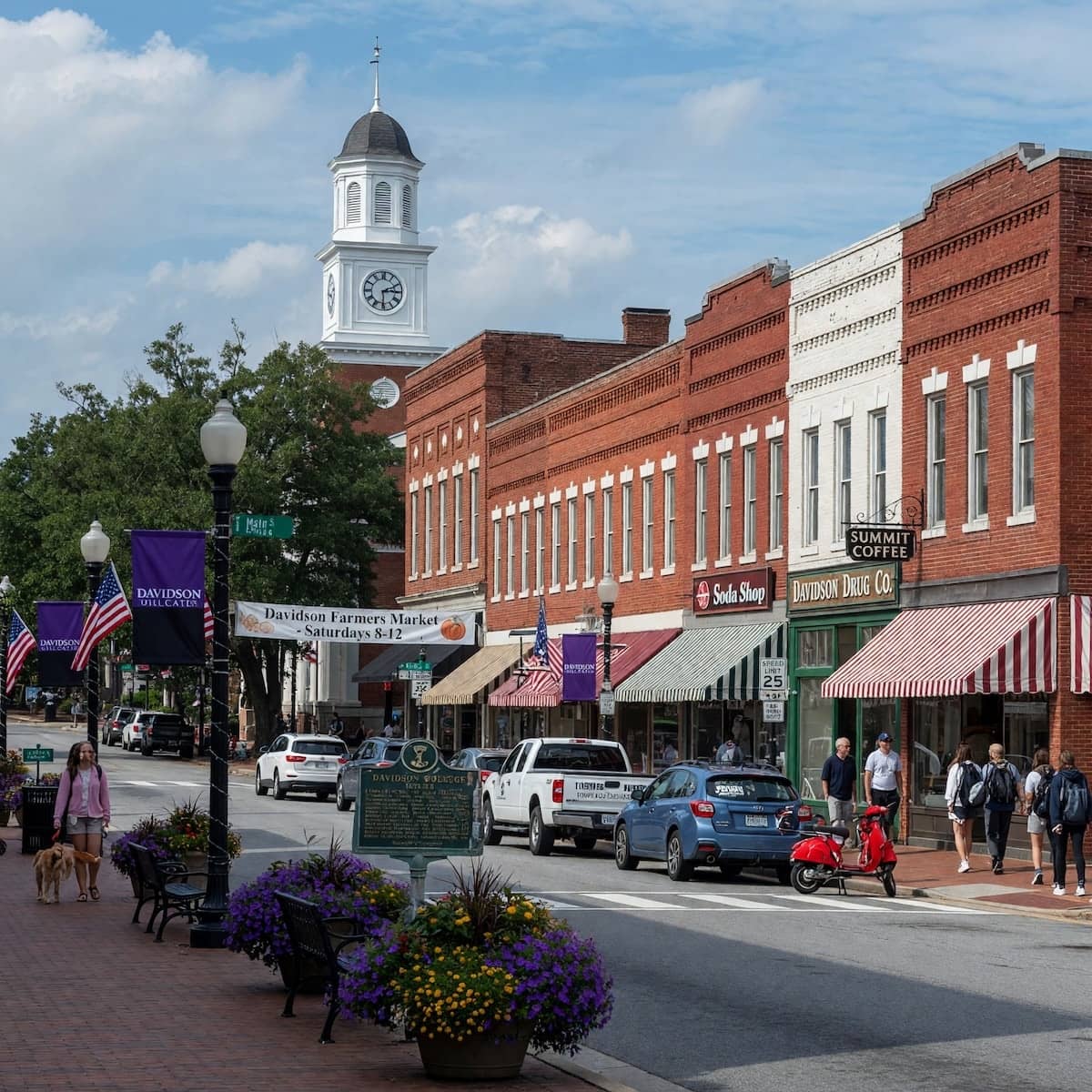A quaint street scene in downtown Davidson, North Carolina, featuring historic red-brick storefronts with striped awnings, flower planters along the sidewalk, pedestrians walking, and a tall white clock tower in the background.