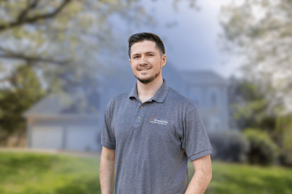Professional home inspector wearing a grey collared shirt standing outdoors in front of a residential house.