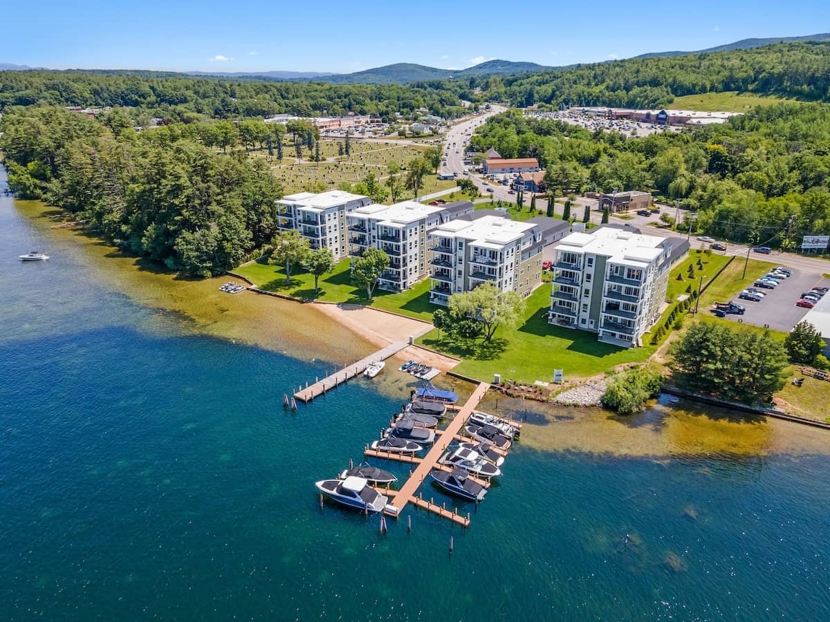 An aerial view of modern lakefront condominium buildings with a private boat dock along clear blue water, surrounded by lawns and trees.