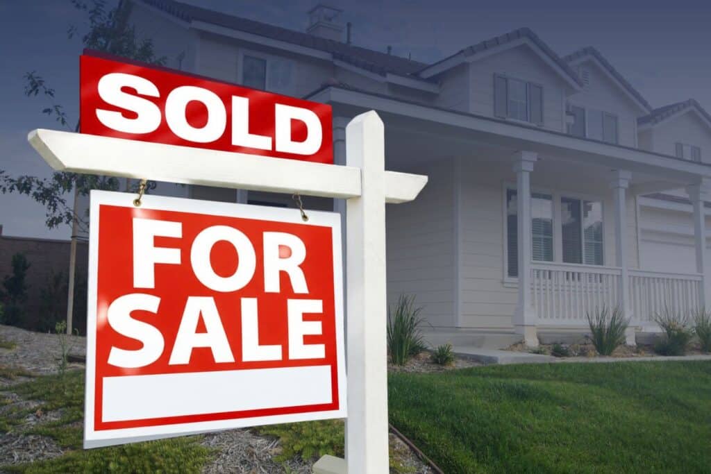 A red and white real estate sign with a "SOLD" banner attached above a "FOR SALE" sign in front of a suburban home.