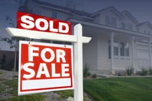 A red and white real estate sign with a "SOLD" banner attached above a "FOR SALE" sign in front of a suburban home.