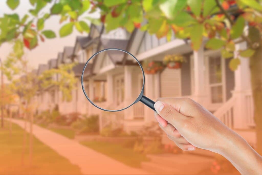 A hand holding a magnifying glass up toward a suburban house in a residential neighborhood.